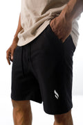 A person wearing black shorts, viewed from the side with one hand on the pocket, showing a small white Gym stereo logo on the leg.